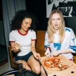 Dwie młode kobiety jedzą pizzę na żółtej kanapie podczas domowej imprezy