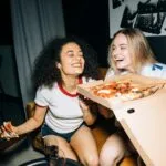 Dwie przyjaciółki jedzą pizzę i śmieją się podczas wieczoru w domu